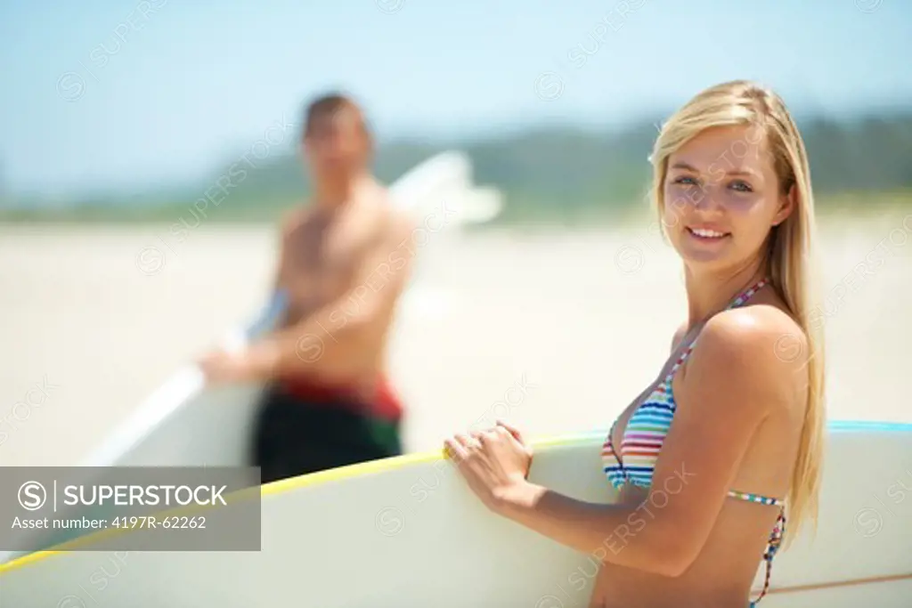 A pretty surfer standing on a beach carrying a surfboard under her arm with fellow surfer standing in the background