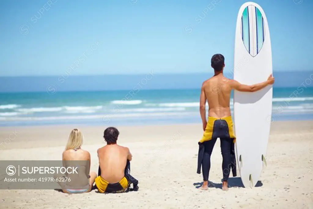 A couple sitting on a beach looking at the ocean with a friend standing next to them holding a surfboard