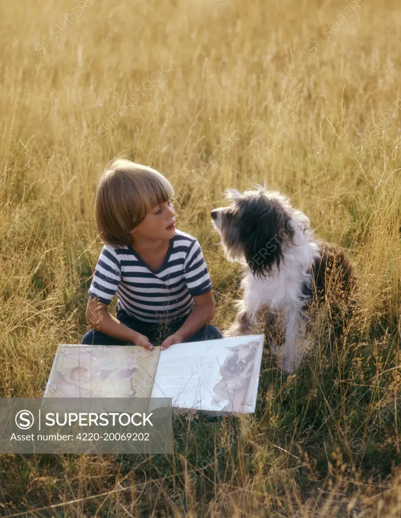 Young boy reading with his dog, a Bearded Collie. Richmond Park, London, UK.