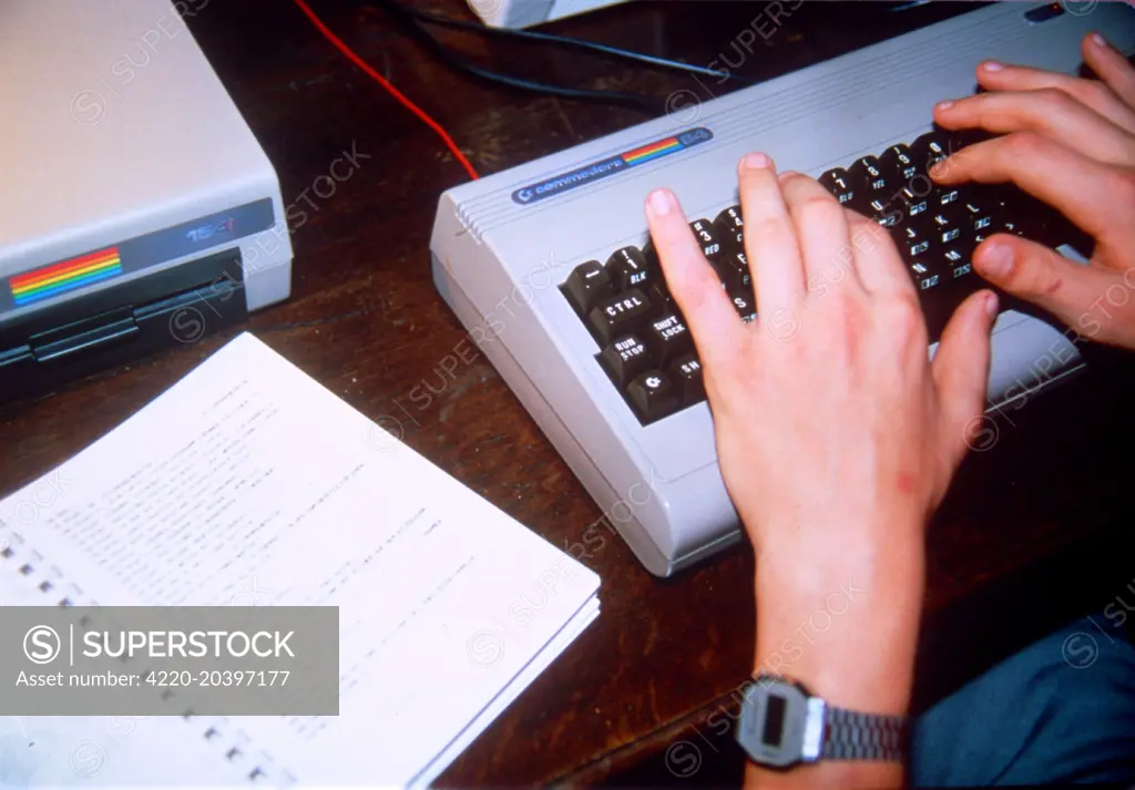 A person wearing a digital  watch types at a Commodore  keyboard. A notebook and  floppy disk drive sit on the  desk beside.      Date: c.1982