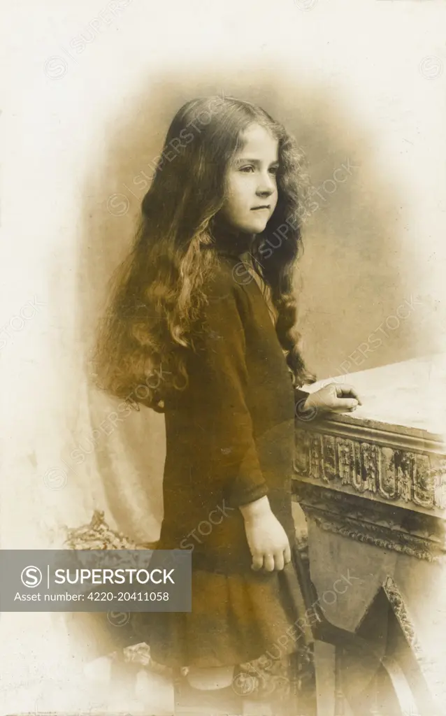 A fabulous photographic postcard of a young girl, barely into her teens with extraordinary long brown, slightly wavy hair, resting against a stone table.     Date: circa 1910s