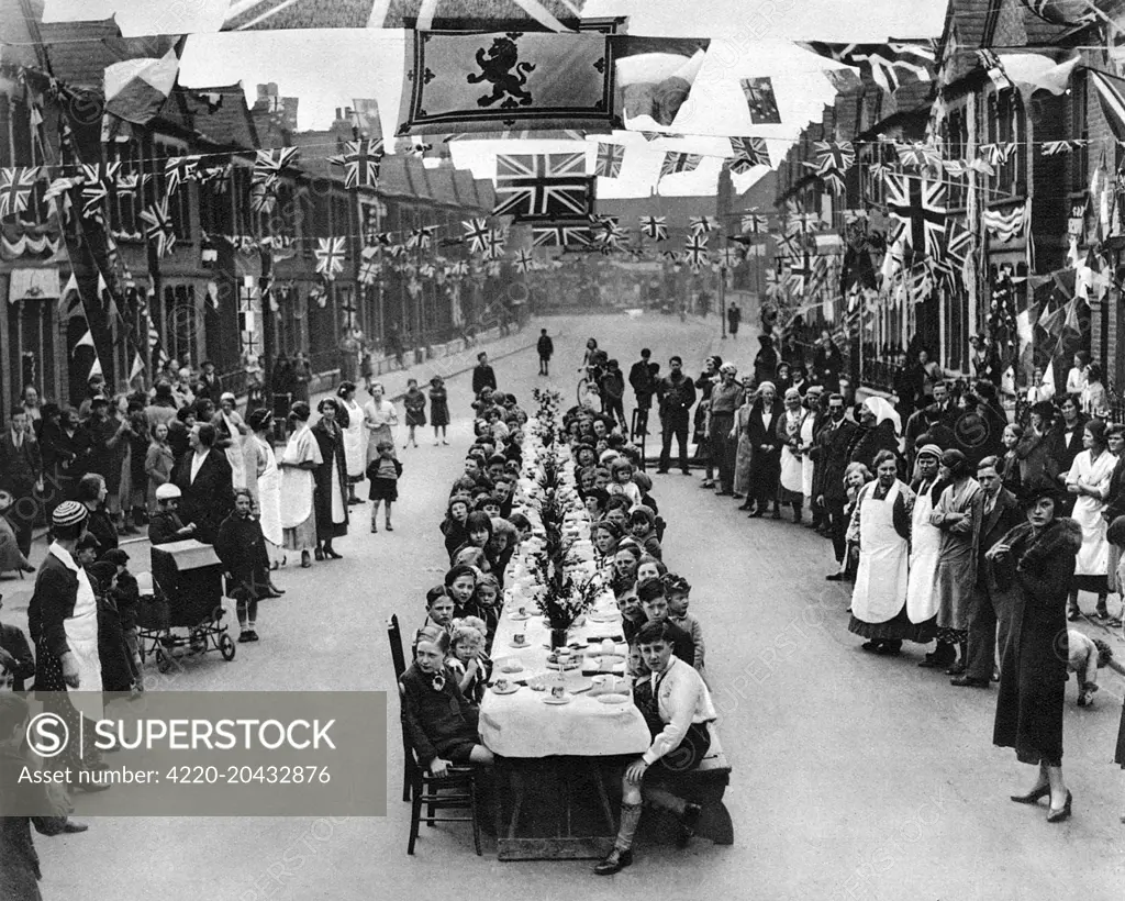A street party in Edenvale Road, Fulham, London to celebrate the Coronation of Kinlocal children seated at a long table for a tea party and flags and bunting festooned across the street.     Date: 1937