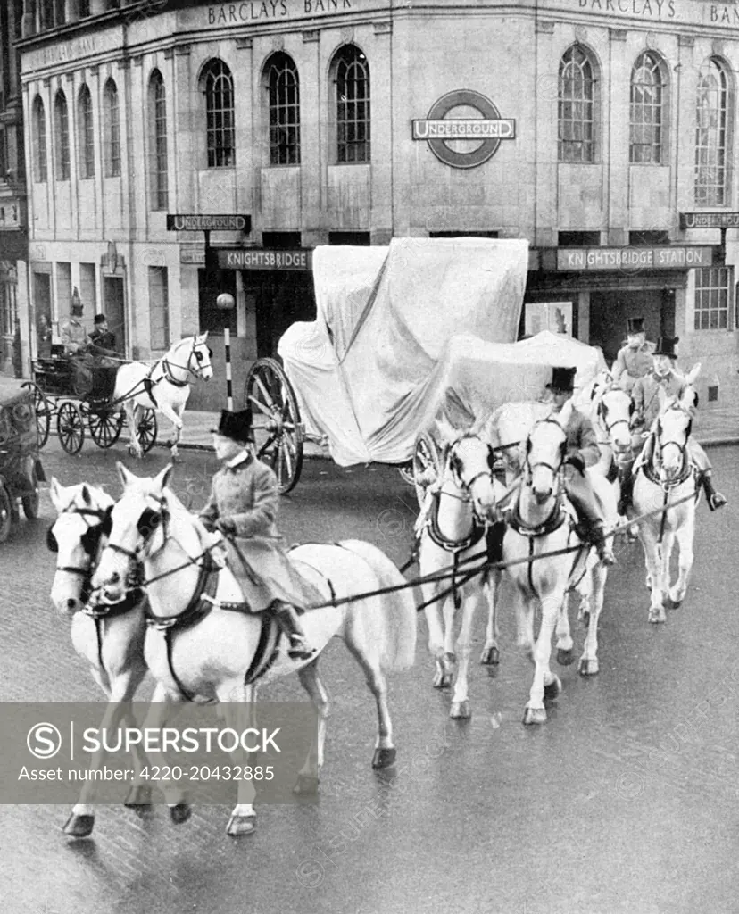 The 4 ton golden Coronation (state) coach is pulled by the famous Royal Greys through Knightsbridge shrouded in cloth, returning to Buckingham Palace after being renovated by a firm of Chelsea coach builders prior to the 1937 Coronation of King George VI.  The coach was first used at an opening of Parliament in the reign of King George III and has been used for every Coronation since that of King George IV.       Date: 1937
