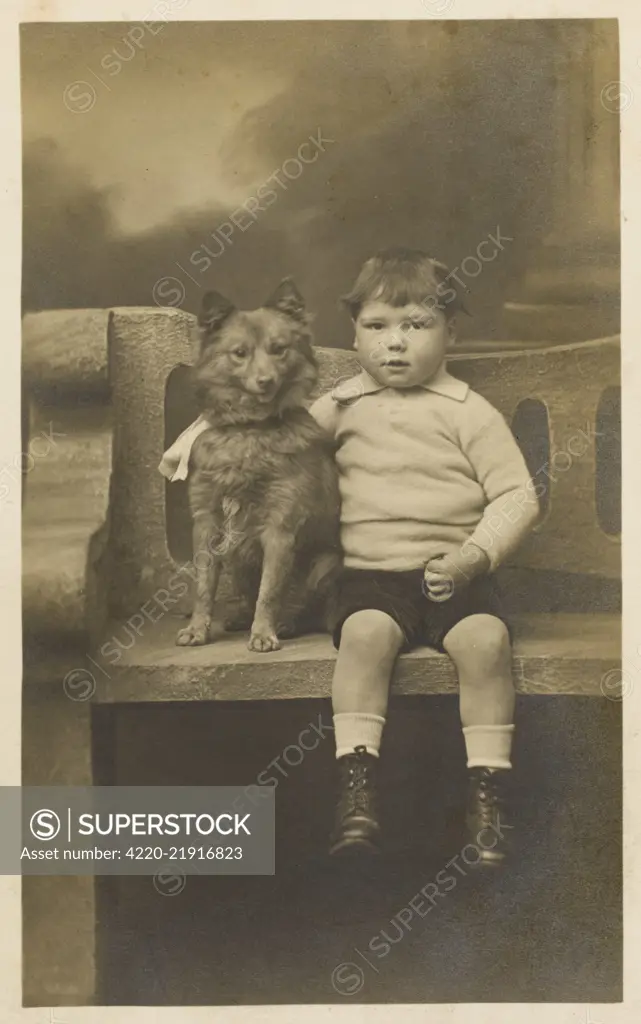 A chubby young child with a  pudding bowl haircut poses for  a studio portrait with his pet  Pomeranian.       Date: Early 20th century
