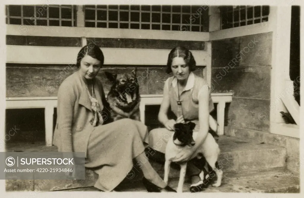 Two women with dogs (an Alsatian and a terrier) in a back garden.     Date: circa 1930s