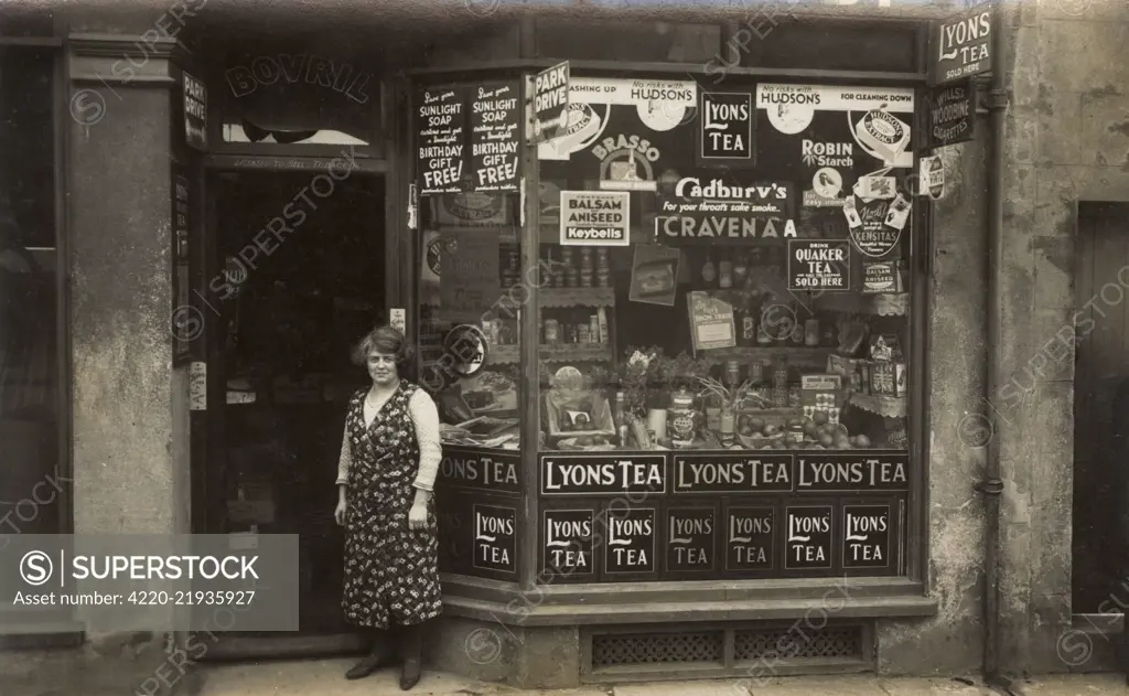Woman standing outside a grocer's shop, with window display and numerous advertisements.      Date: circa 1930s