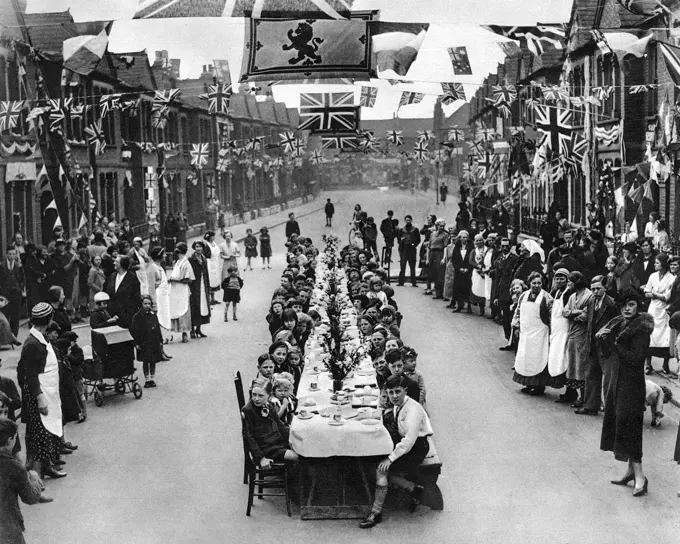 A street party in Edenvale Road, Fulham, London to celebrate the Coronation of Kinlocal children seated at a long table for a tea party and flags and bunting festooned across the street.     Date: 1937