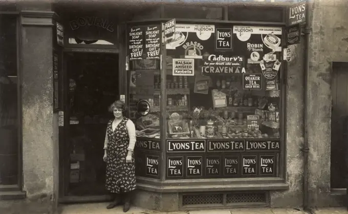 Woman standing outside a grocer's shop, with window display and numerous advertisements.      Date: circa 1930s