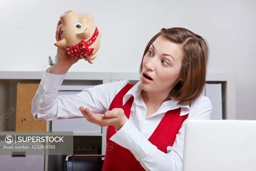Shocked woman in office shaking a piggy bank