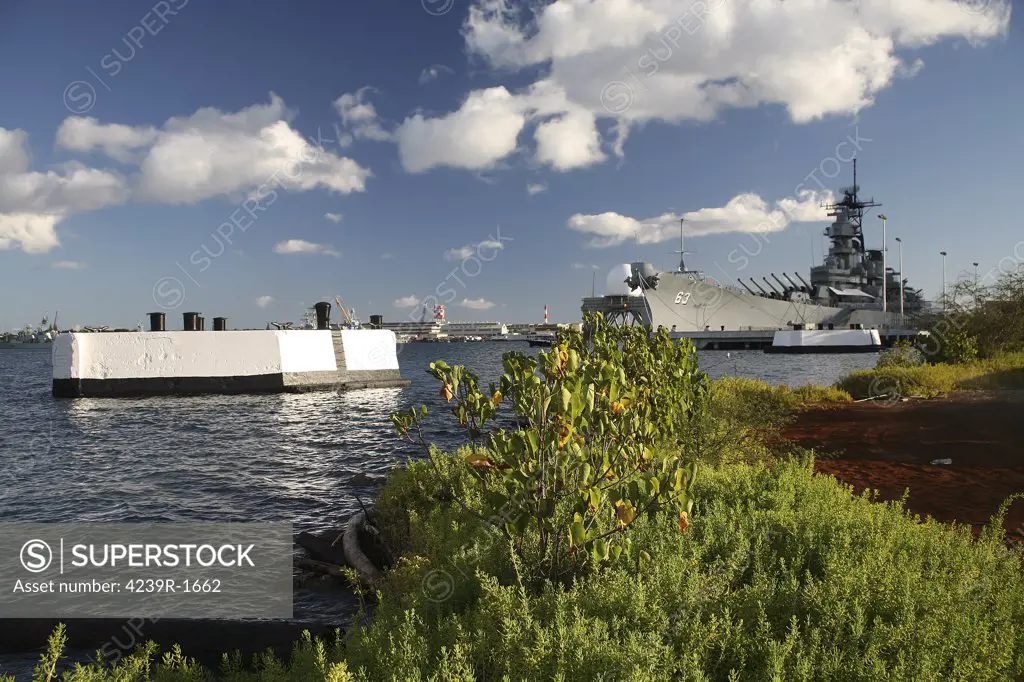 A battleship row mooring pier and Ford Island plant growth frame the foreground of the USS Missouri, BB-63, Mighty Mo or Big Mo, as it is berthed pierside at Ford Island, Oahu, Hawaii