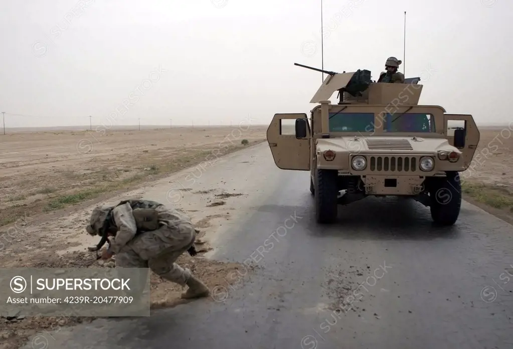 U.S. Marine checks a pothole for mines on Main Supply Route Tin, during a convoy from Camp Al Asad to Al Qaim, during Operation Iraqi Freedom.  The USMC M1116 Up-Armored High-Mobility Multipurpose Wheeled Vehicle (HMMWV) armed with a Browning M2 HB 0.50 caliber heavy machine gun on a turret waits for the all clear.