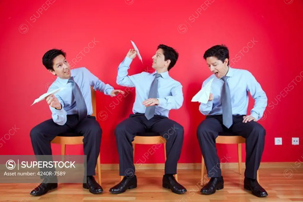 Three Twin Businessman Sited Next To A Red Wall Trowing Paper Planesl