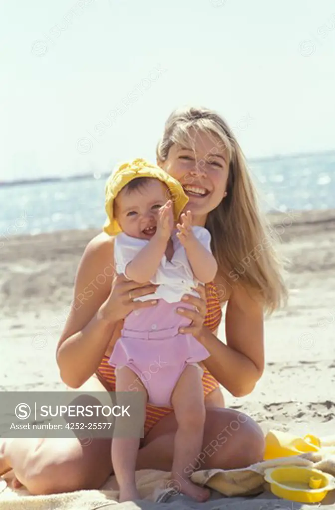 family outside sea woman baby sun protection summer mother mummy parent embracing child suntan beach sand sitting hat swimsuit dress t-shirt complicity look applaud movement hand stand up smiling laughing holidays happiness