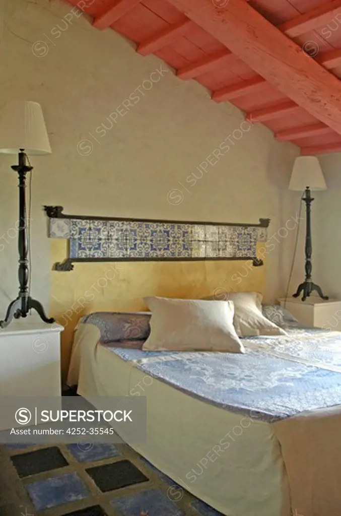 Oriental styled attic bedroom with wooden beams painted into pink, tiled grey and blue floor, bedhead pannel on the wall made of oriental tiles and tall lamps on the bedsides