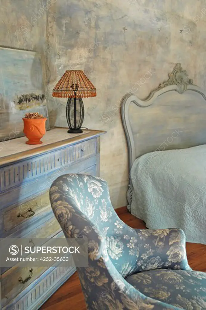 Blue flowered armchair into a bedroom, with a chest of drawers painted with a blue patina