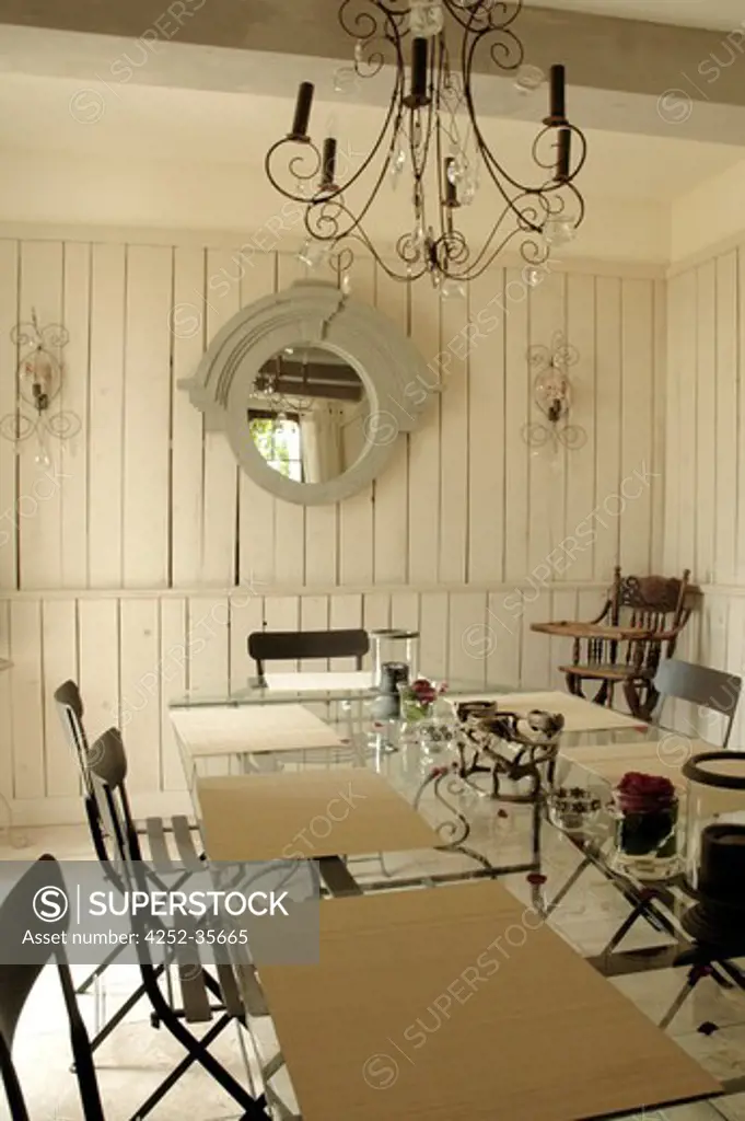 White dining room with a large glass table, a chandelier hanging on the ceiling and wooden white walls