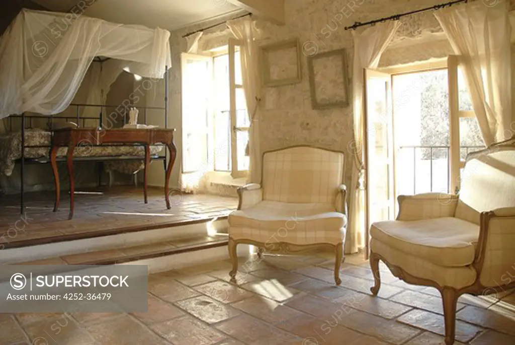 Old armchairs into a beige bedroom with a canopied bed on the back and a tiled floor