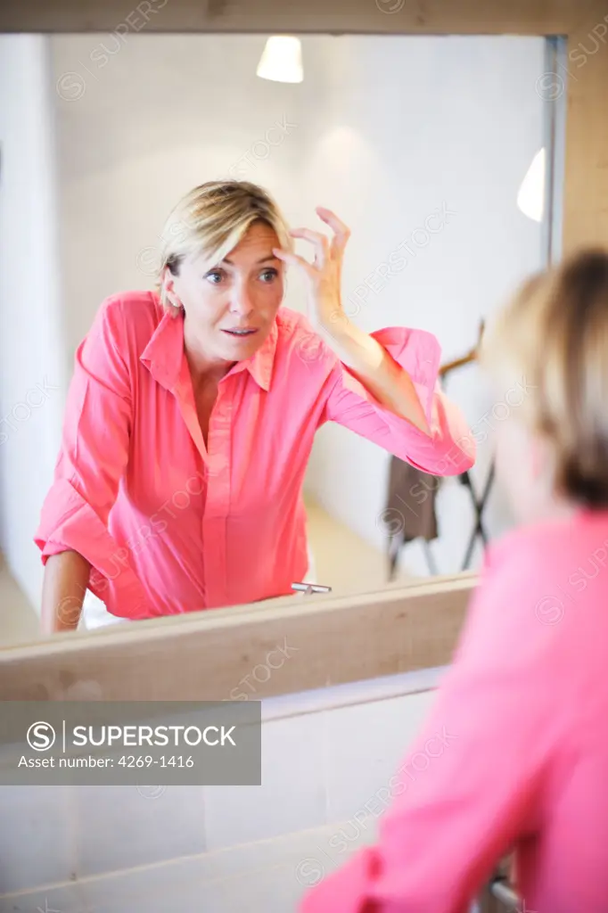 Woman checking her face for wrinkles in the mirror. Wrinkles occur when skin loses its elasticity and suppleness as it ages.