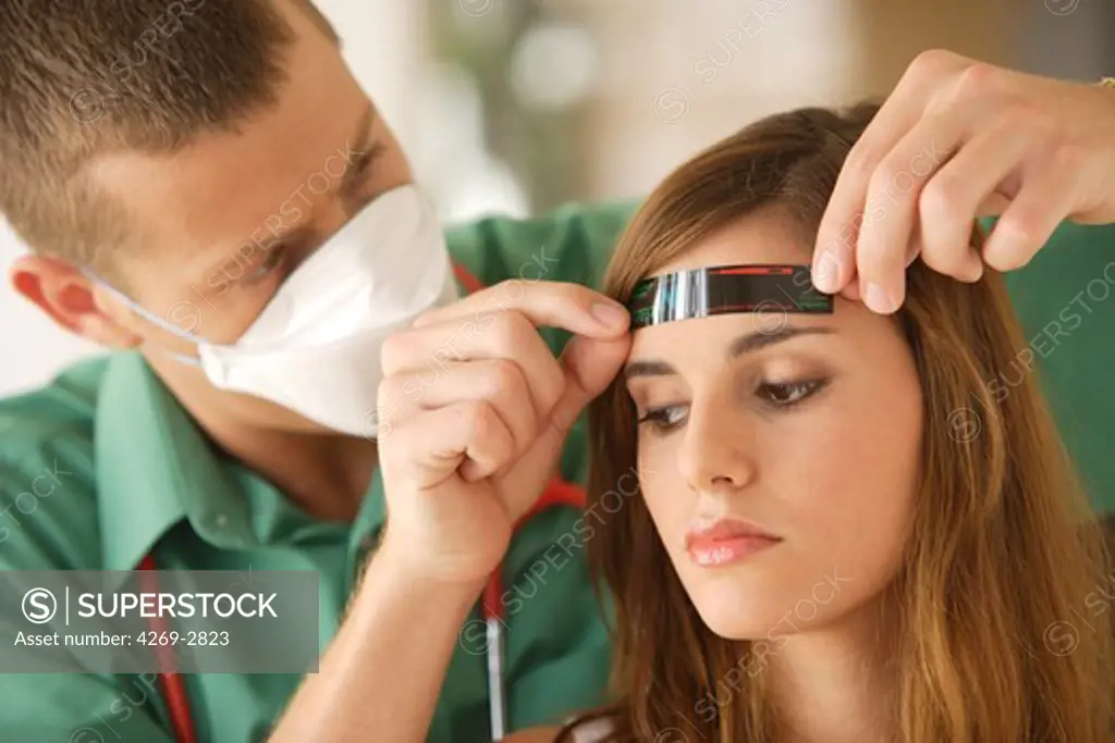 General practitioner wearing a respiratory protection mask during consultation with contagion hazard, checking the temperature of a patient with a strip thermometer.