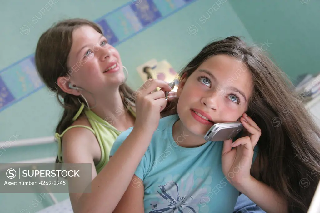12 years old girl listening to music on mp3 player and her 11 years old sister talking on cell phone.
