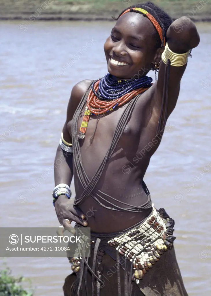 A young Dassanech girl beside the Omo River.  Her hairstyle, necklaces and metal armbands are typical of her tribe.  The broad belt attached to her leather skirt is decorated with seeds and hundreds of tiny ostrich eggshell discs one of the earliest forms of personal adornment in Africa, which dates back thousands of years.