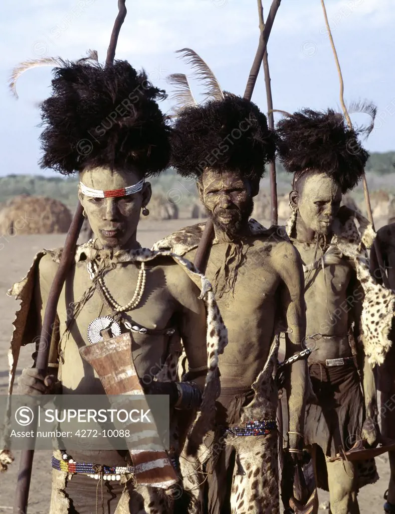Men dance during a month long Dassanech ceremony.  They wear leopard, cheetah or serval cat skins draped on their backs and black ostrich-feather headdresses.  They dance holding long sticks and simulated shields. Their faces and bodies are smeared with mud giving them a singular appearance.
