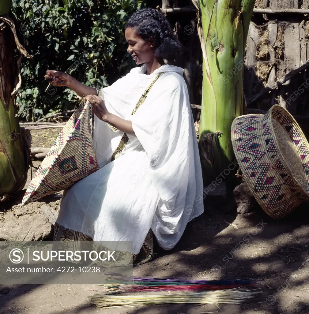 A young Amhara lady weaves a traditional food basket from dried grasses. These large colourful baskets are used for serving injera, a fermented, bread-type pancake, which is the country's national dish.She is wearing the national dress of Ethiopia - a shamma. This garment is made of homespun cotton with a finely woven and often brightly coloured border.