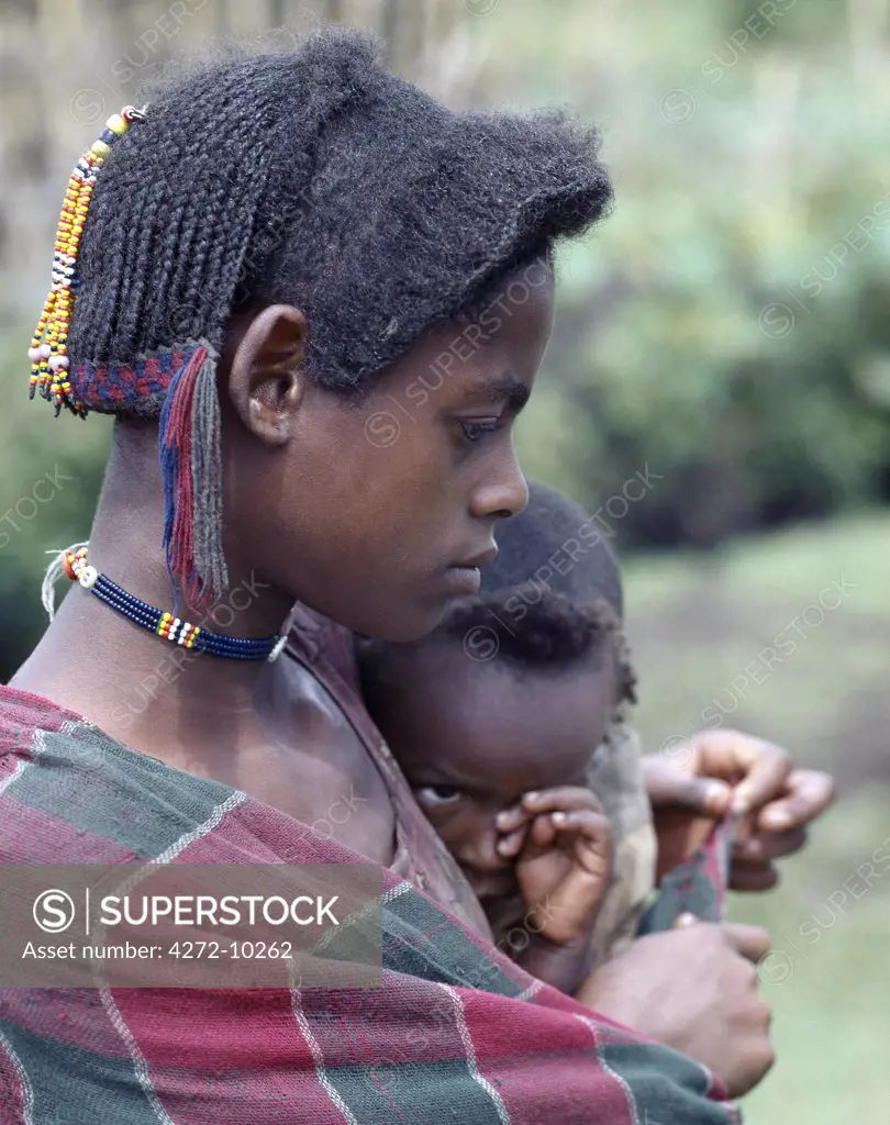A young mother and child of the Arsi-Oromo people west of Aje.  Both have unusual hairstyles. The braids falling from the crown of the mother's head have been attractively woven with wool to make a colourful fringe.