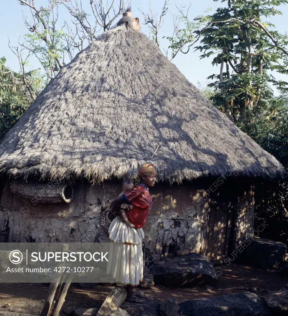 A mother and child outside a Konso house in southwest Ethiopia. All the high pitched, thatched roofs of Konso houses are topped with earthenware pots to keep out rain. Hanging beneath the eaves of this house is a traditional cylindrical beehive.The womans skirt is typical of the style worn by Konso women and is made from thick cotton cloth, called buluko, weaved locally by men.