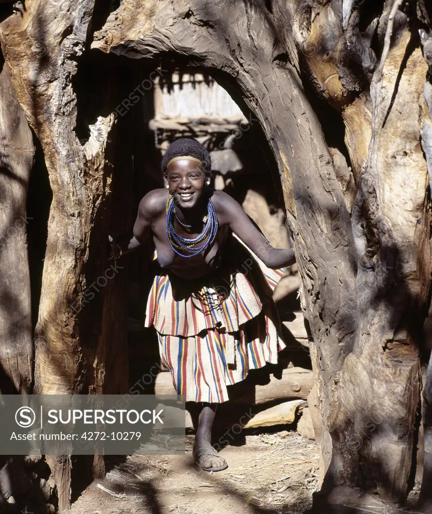 A young woman at the entrance to a Konso homestead in southwest Ethiopia. The konso have a great affinity for wood and stone; large tree trunks and branches surround every home, and special care is taken to select the most pleasing shapes for their entrances.The womans skirt is typical of the style worn by Konso women and is made from thick cotton cloth, called buluko, weaved locally by men.