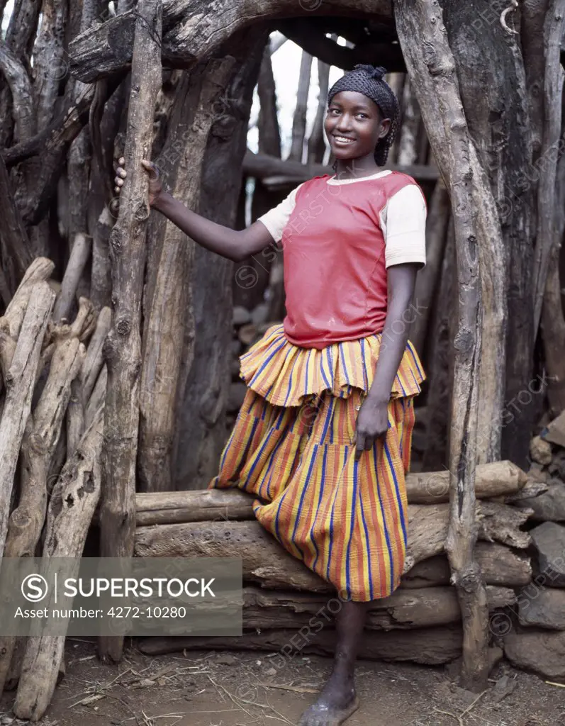 A young woman at the entrance to a Konso homestead in southwest Ethiopia. The konso have a great affinity for wood and stone; large tree trunks and branches surround every home, and special care is taken to select the most pleasing shapes for their entrances. The womans skirt is typical of the style worn by Konso women and is made from thick cotton cloth, called buluko, weaved locally by men.