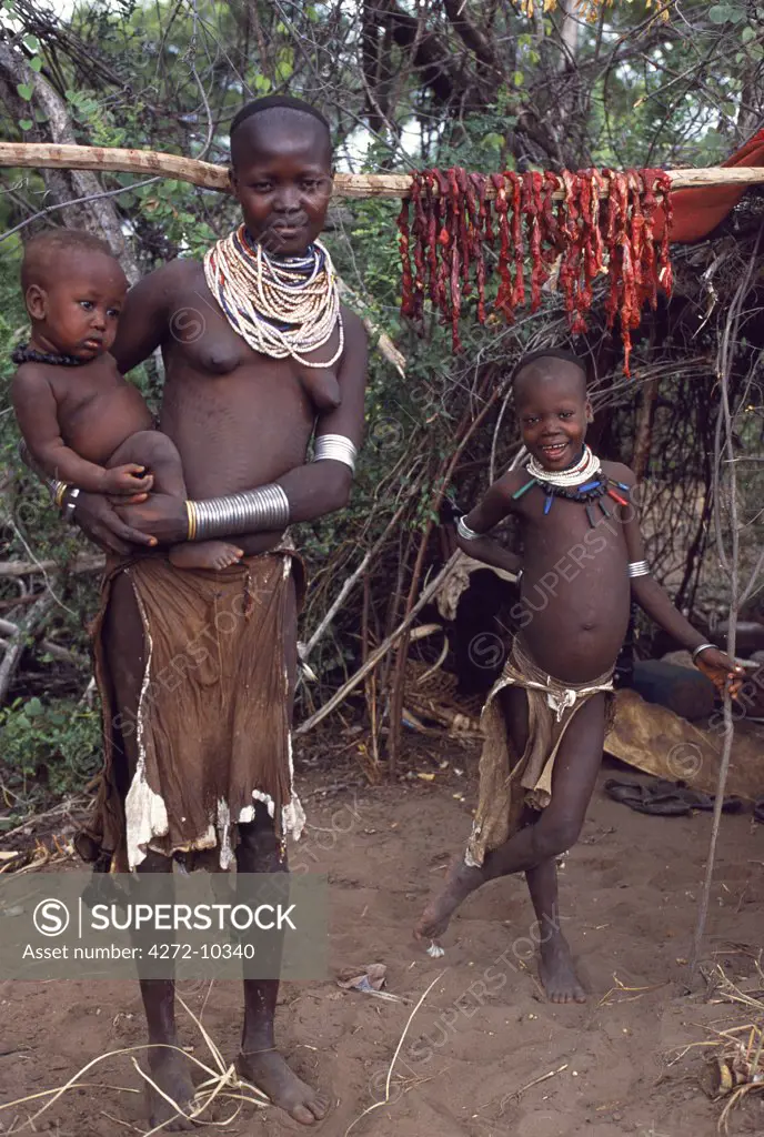 A Karo woman carries her baby on her hip at their temporary camp on the bank of the Karo River.  Meat from a  slaughtered animal hangs drying on a pole outside her hut.  A small Omotic tribe related to the Hamar, who live along the banks of the Omo River in southwestern Ethiopia, the Karo are renowned for their elaborate body painting using white chalk, crushed rock and other natural pigments.