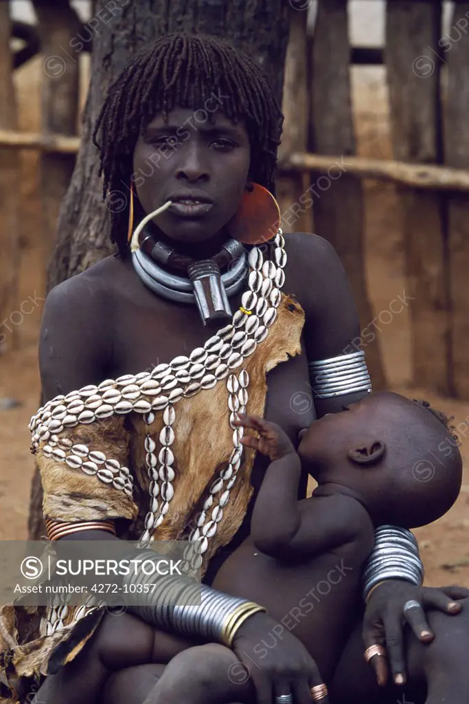 A Hamar woman sits on the ground breast-feeding her baby in the village square of Dimeka.  Married women wear two heavy steel necklaces.  This woman wears an extra necklace with steel a steel phallic symbol which identifies her as a first wife.  She also wears a goatskin capefringed with cowrie shells and her hair long in a braided fringe matted with animal fat and ochre.