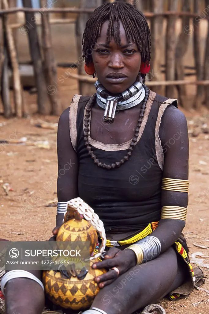 A Hamar woman sits holding a gourd in the village square of Dimeka.  Married women wear two heavy steel necklaces.  This woman wears an extra necklace with steel a steel phallic symbol which identifies her as a first wife.  She wears her hair long in a braided fringe matted with animal fat and ochre.