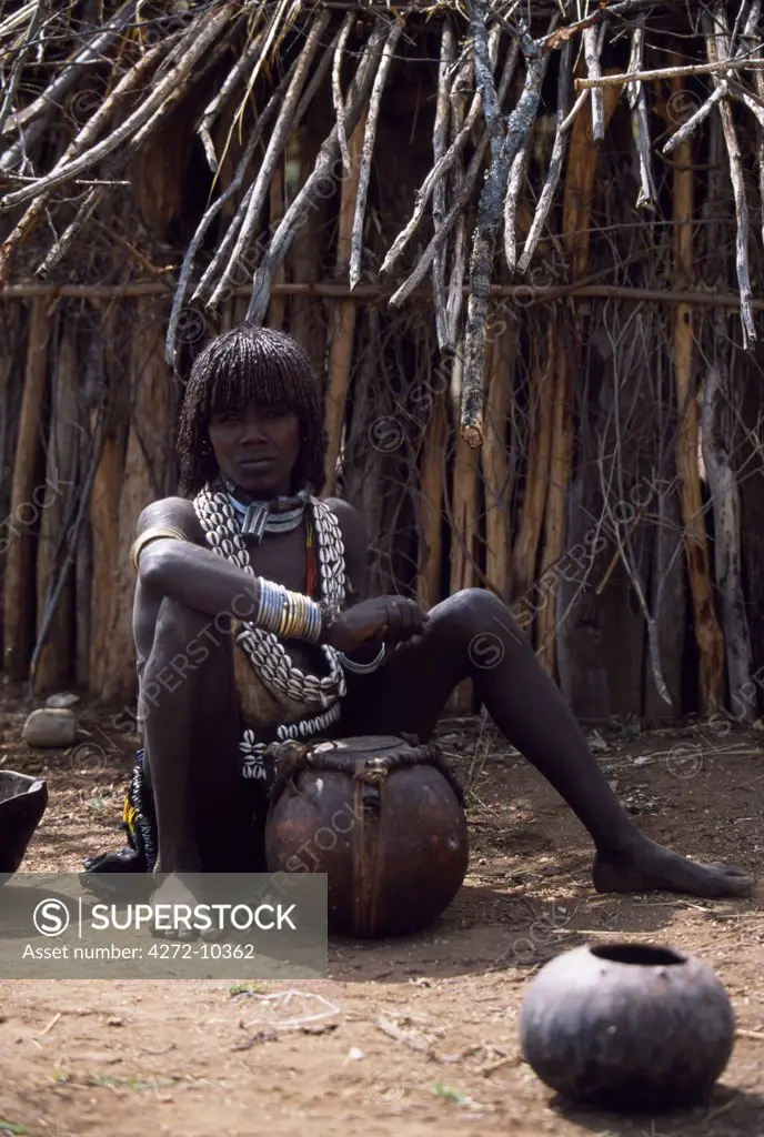 A Hamar woman sits in front of her hut with a gourd of fermented sorghum beer.  She is wearing an intricately beaded leather skirt and apron, decorated with cowrie shells.  Her hair is braided and reddened with a mixture of ochre and animal fat.  She wears the two heavy iron necklaces of a married woman and the extra necklace with phallic symbol that identifies her as a first wife.
