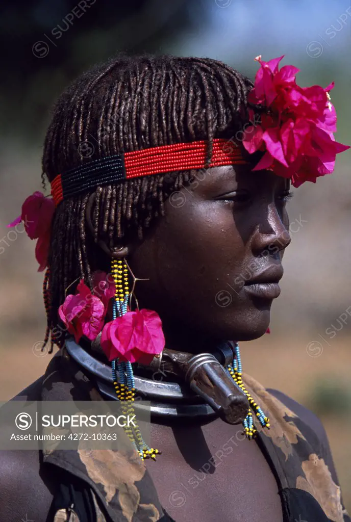 A Hamar woman wears the two heavy iron necklaces of a married woman and the extra necklace with phallic symbol that identifies her as a first wife.  Her hair is braided and reddened with a mixture of ochre and animal fat and decorated with bougainvillia flowers.