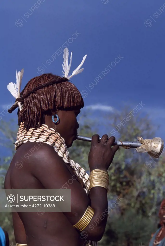 A Hamar woman blows a tin horn at the bull -jumping ceremony of a man in her family, a rite of passage to manhood.  She wears a leather belt decorated with cowrie shells around her neck.  As is normal for her tribe she wears her hair braided and thick with ochre and animal fat.