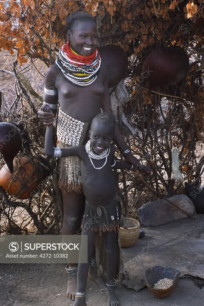 A Nyangatom woman and child stand by her temporary shelter.  The adult women wear many layers of bead necklaces and elegant skirts with a short beaded fringe at the font and long calfskin tail reaching the ground at the back. The Nyangatom or Bume are a Nilotic tribe of semi nomadic pastoralists who live along the banks of the Omo River in south western Ethiopia.