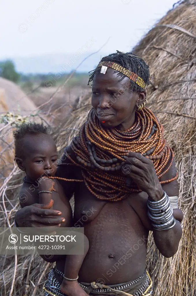 A Nyangatom woman stands with her baby on her hip beside her grass hut in his temporary camp.  Nyangatom married women wear elaborately beaded skirts which reach the ground at the back and often have panels of different coloured calkfskin sewn into the tail, many layers of bead necklaces and iron bracelets, amulets and anklets.