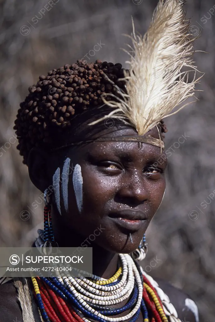 A young Nyangatom woman has painted her face and body and put a feather in her hair in preparation for a dance.  Her hair has been reddened with a mixture of ochre and animal fat.  She has a pierced lower lip through which she has inserted a nail and she is wearing a goatskin apron.  The Nyangatom or Bume are a Nilotic tribe of semi-nomadic pastoralists who live along the banks of the Omo River in south-western Ethiopia.