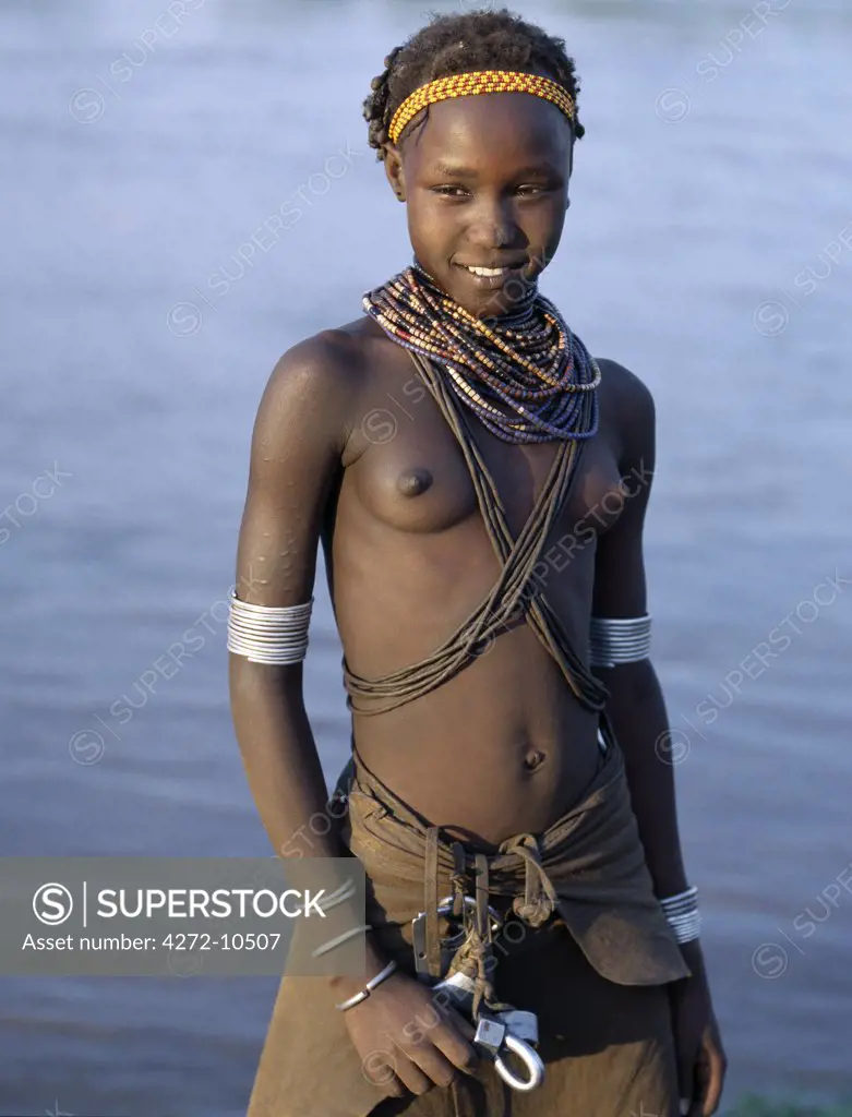 An attractive Dassanech girl stands on the banks of the Omo River. Her leather skirt and adornment are typical of the girls of her tribe. Since the Omo Delta is one of the least accessible and least developed parts of East Africa the culture, social organization, customs and values of the people have changed less than elsewhere.