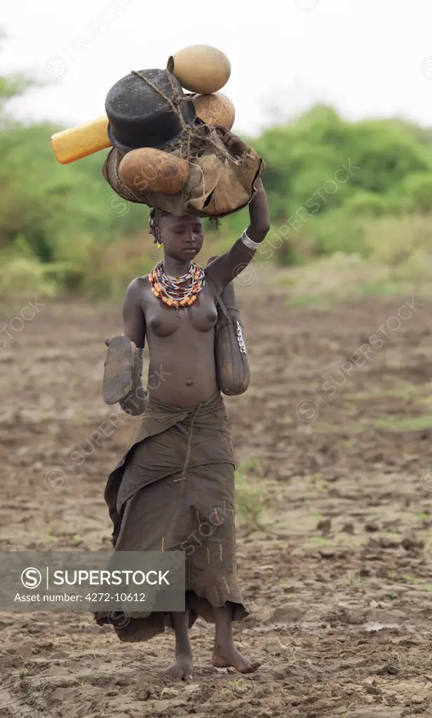 A Dassanech girl with sandals made from an outer tyre in her hand walks through muddy soil along the banks of the Omo River carrying a head load of cooking pots and gourds. The Dassanech speak a language of Eastern Cushitic origin. They live in the Omo Delta and they practice animal husbandry and fishing as well as agriculture.