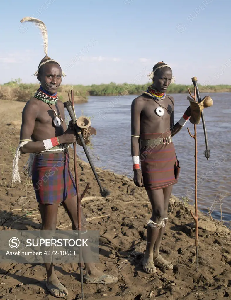 Two smartly dressed Dassanech men stand on a bank of the Omo River. Their attire signifies that they are about to marry.  Note the oryx horns they are carrying in which they keep their ostrich feathers for adorning their clay hairdos.The Omo Delta of southwest Ethiopia is one of the least accessible and least developed parts of East Africa.