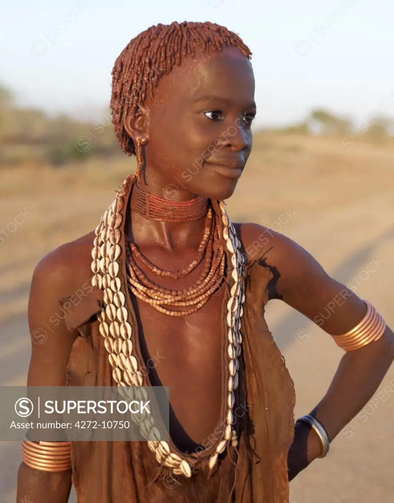 The ochre worn by a pretty young Hamar girl glows deep red in the late afternoon light.;The Hamar are semi-nomadic pastoralists of Southwest Ethiopia whose women and girls wear striking traditional dress. Skins are widely used for clothing and heavy metal necklaces, bracelets and anklets form part of a woman's adornments. Cowries are also popular to embellish their appearance.