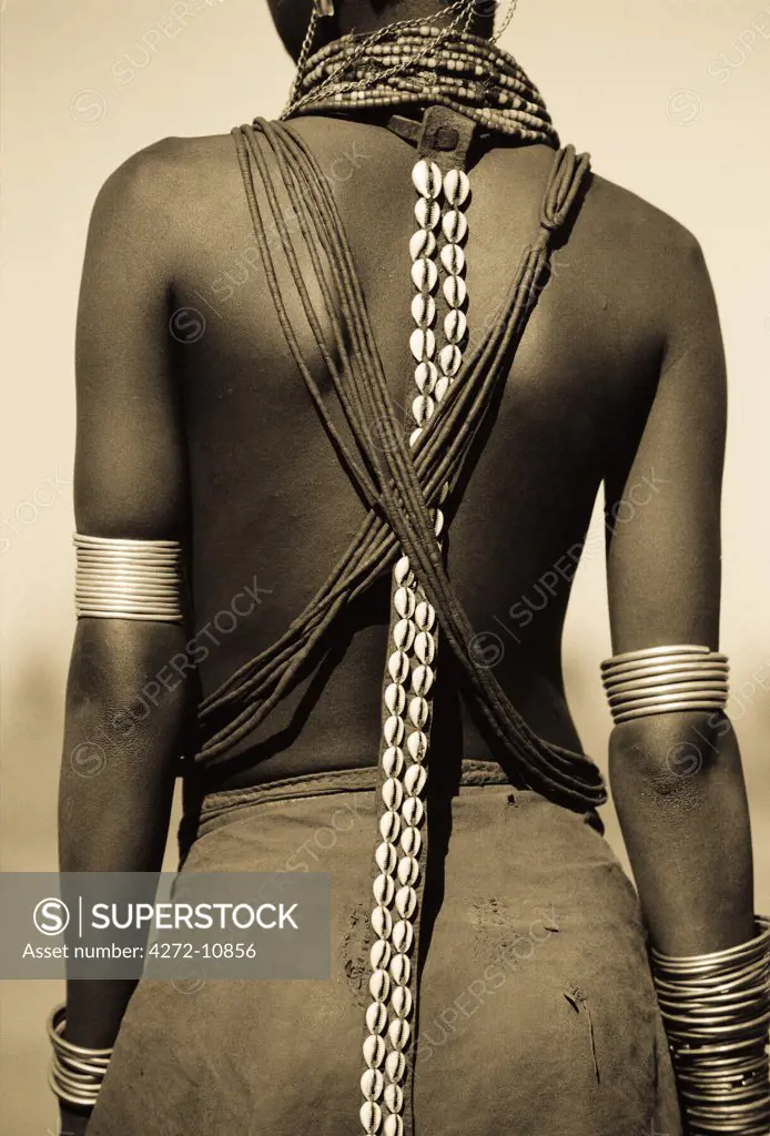 Ethiopia, Omo Delta.  A young Dassanech girl wears a leather skirt, metal bracelets and amulets and beadss. A leather strap with cowrie shells hangs down her back.