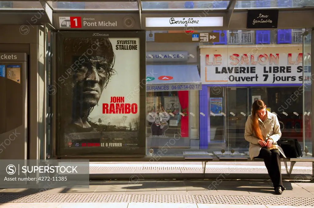 France, Cote D'Azur, Nice; A girl waitng at one of the tram stations in Avenue Jean Medecin with a poster advertising a rambo film
