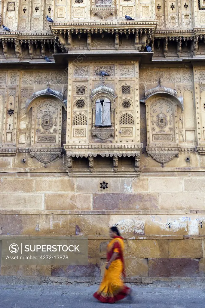 India, Rajasthan, Jaisalmer. On the outside walls of the city's largest haveli, street life goes on as normal as a sari clad woman walks alongside the heavily decorated walls.