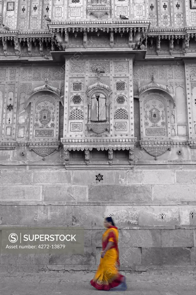 India, Rajasthan, Jaisalmer.  On the outside walls of the city's largest haveli, street life goes on as normal as a sari clad woman walks alongside the heavily decorated walls.