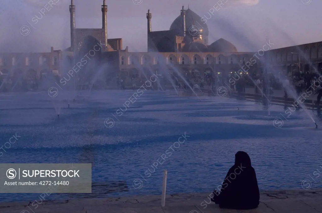 Iran, Isfahan. A woman in traditional dress sits looking at  the fountains of Imam Khomeini Square
