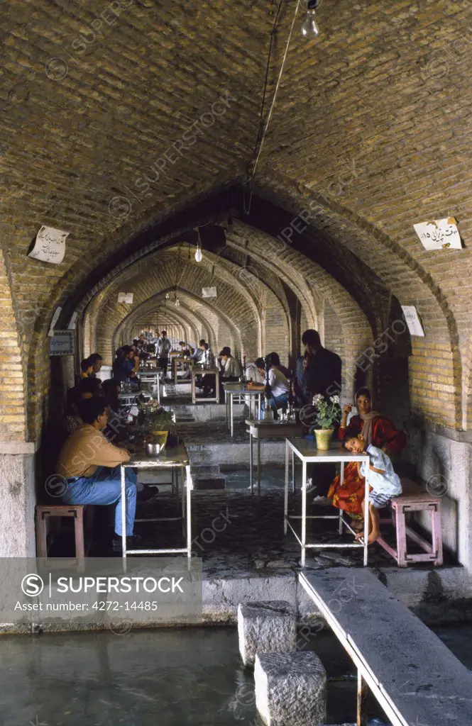 Iran, Esfahan. Iranians sit drinking tea at tables in a tea house beneath the arches of one of Esfahan's bridges across the Zayande River